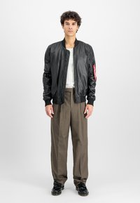 Young man standing, wearing black leather bomber jacket, white shirt, brown wide-leg trousers, and black shoes against a plain white background.