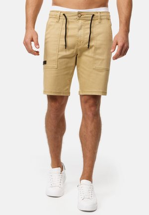 Beige Cargo-Shorts aus weicher Baumwolle. Verfügen über zwei Fronttaschen, einen verstellbaren Kordelzug und ein schwarzes Etikett am linken Oberschenkel. Kombiniert mit weißen Schuhen.