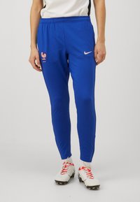 Mėlyni atletiniai džinsai su aptemptu siluetu, kuriuose matomas rožinis Nike logotipas ir gaidžio emblema kairėje šlaunyje. Pagaminti iš glotnaus, lengvo audinio.