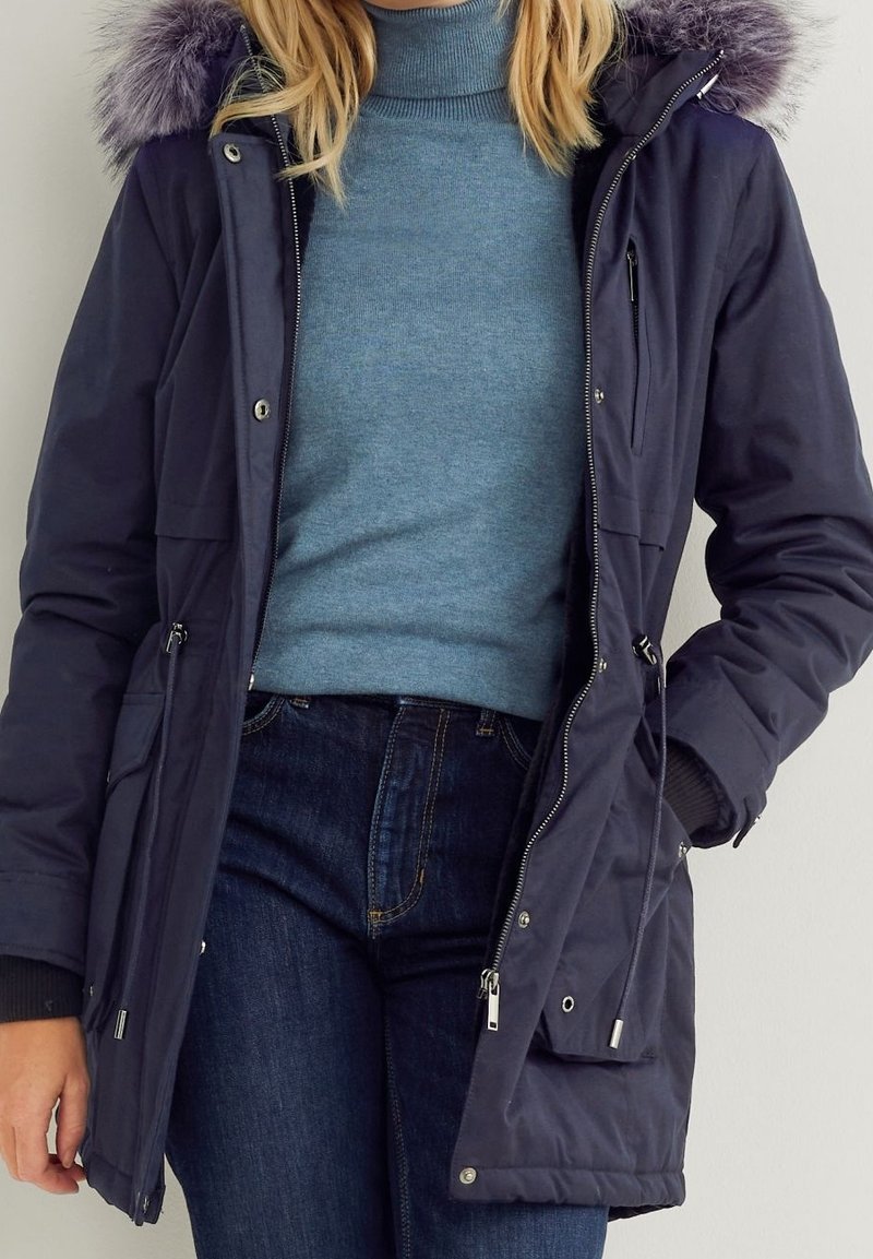Parka bleu marine avec capuche bordée de fausse fourrure, fermeture éclair à l'avant, poches latérales et poignets côtelés, portée sur un col roulé bleu clair.