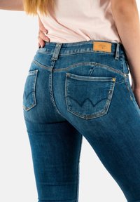 Jean en denim bleu ajustés avec une taille mi-haute, des coutures distinctives sur les poches arrière et un patch en cuir sur la ceinture.