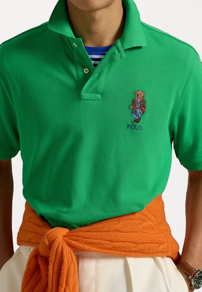 Roheline polo särk tekstuuriga viimistlusega, millel on tikitud karu disain ja "POLO" tekst. Oranž soonikkoes kampsun on seotud vöökohtadele.