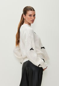 Suéter blanco de punto con patrón de trenzas, con detalles de cintas negras en las mangas. Combinado con elegantes pantalones de cuero negros.