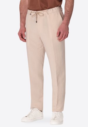Uomo con pantaloni beige con coulisse, sneaker bianche e camicia marrone, in piedi davanti a uno sfondo chiaro e uniforme.