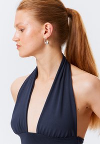 Top blu navy con scollatura a halter, scollatura profonda, tessuto morbido e dettaglio arricciato in vita. Orecchino a cerchio argentato completa il design.