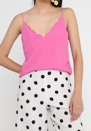 Femme portant un caraco rose à bord festonné et un pantalon blanc à gros pois noirs, debout avec une main sur la hanche.