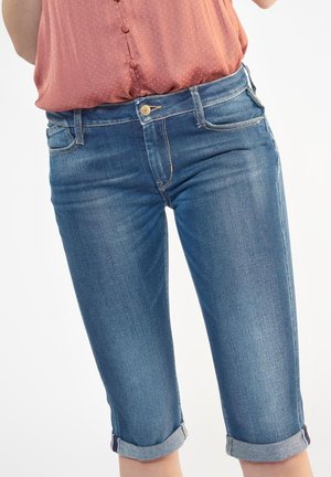 Femme portant un pantacourt en denim bleu retroussé et un chemisier rose boutonné rentré, visible de la taille jusqu'aux genoux.