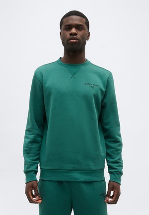 Fiatal férfi zöld Björn Borg pulóverben és hozzáillő nadrágban, sima világosszürke háttér előtt áll.