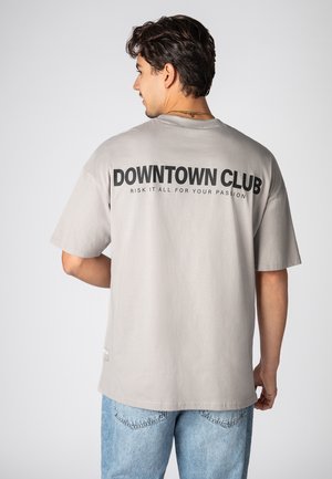 Helles graues Baumwoll-T-Shirt mit kurzen Ärmeln, das auf der Rückseite großen schwarzen Drucktext mit der Aufschrift "DOWNTOWN CLUB" und einem kleineren Slogan darunter zeigt.