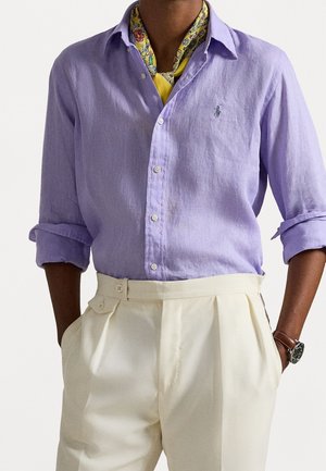 Homme portant une chemise lavande à boutons avec manches retroussées, un foulard jaune à motifs autour du cou, un pantalon plissé crème taille haute et une montre-bracelet.