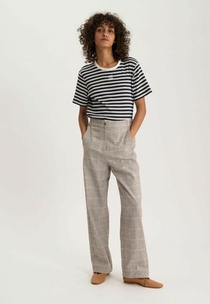 Femme aux cheveux bouclés, debout les mains dans les poches, portant un t-shirt rayé noir et blanc, un pantalon à carreaux beige et des chaussures plates couleur tan.