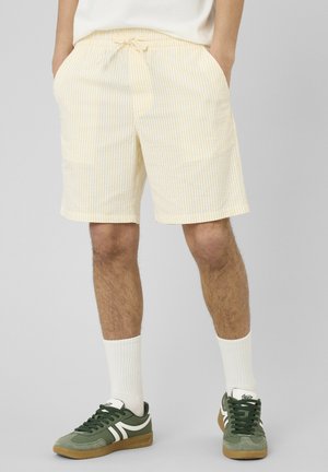 Homme portant un short rayé jaune clair et blanc avec des poches, des chaussettes blanches et des baskets vertes à semelles en gomme, debout devant un fond uni.