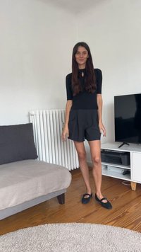 Čierny priliehavý top s otvorom, doplnený čiernymi šortkami s veľkými vreckami a tmavými plochými topánkami. Drevená podlaha a minimal decor.