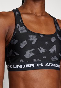 Melnā sporta krūšturis ar pelēkiem abstraktiem rakstiem, ar platu melnu joslu un baltu "UNDER ARMOUR" logotipu, izgatavots no elastīga materiāla.
