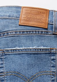 Jeans de mezclilla azul con una etiqueta de cuero marrón cosida encima de la cintura, que presenta un logotipo en relieve y detalles. Costuras dobles y textura deslavada.