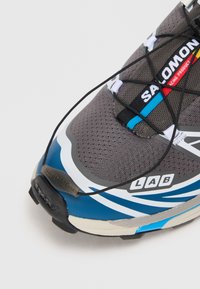 Podrobni posnetek sivo-modre tekaške čevlje Salomon s črnim hitrim vezalnim sistemom in vidnimi logotipi blagovne znamke na jezikih in strani.