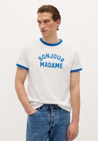 Camiseta blanca de manga corta con cuello y puños de canalé azul. El texto frontal dice "BONJOUR MADAME" en letras azules en negrita. Llevada con vaqueros azules.