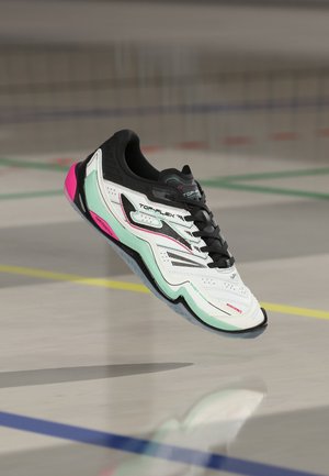 Chaussure de sport d’intérieur blanche et noire avec des touches vert menthe et rose flottant au-dessus d’un sol de gym avec des lignes de terrain colorées.