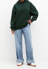 Personne portant un sweat à capuche oversize vert foncé, un jean large bleu clair retroussé et des baskets blanches à rayures bleues, debout contre un mur blanc.
