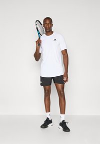 Hvid atletisk t-shirt med sort logo, sorte shorts med hvidt logo, sorte sneaks, holder en tennisracket, mod en ensfarvet baggrund.