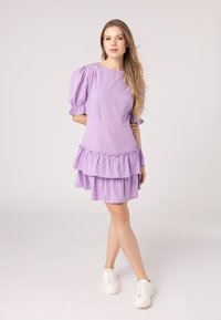 Vestito corto lilla con maniche a palloncino, dettagli a rouches sovrapposti e una vestibilità rilassata. Indossato con sneaker bianche; texture del tessuto liscia.