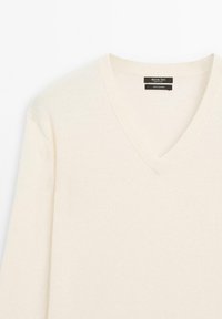 Maglione in cashmere crema con scollo a V e una texture morbida. Presenta dettagli a costine sul colletto e sui bordi. L'etichetta riporta "100% cashmere."