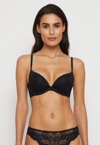Soutien-gorge en dentelle noire avec un motif floral, bonnets rembourrés, bretelles réglables et un petit nœud au centre. Culottes en dentelle assorties visibles.