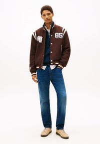 Smeđa akademska jakna s bijelim prugama i vezom, slojevito nošena preko tamnoplave pletene veste i bijele košulje, uparena s plavim trapericama i bež tenisicama.