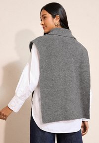 Hallide kudum vest, millel on kõrge krae, avatud külgedega ja soonikstruktuur, kihtidena valge nööbiga särk, mille varrukad on üles keeratud.