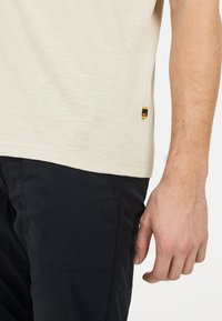 Beige Kurzarmhemd aus strukturiertem Stoff. Mit einem seitlichen Etikett in Schwarz und Gelb. Tragen mit dunkelblauen Hose.