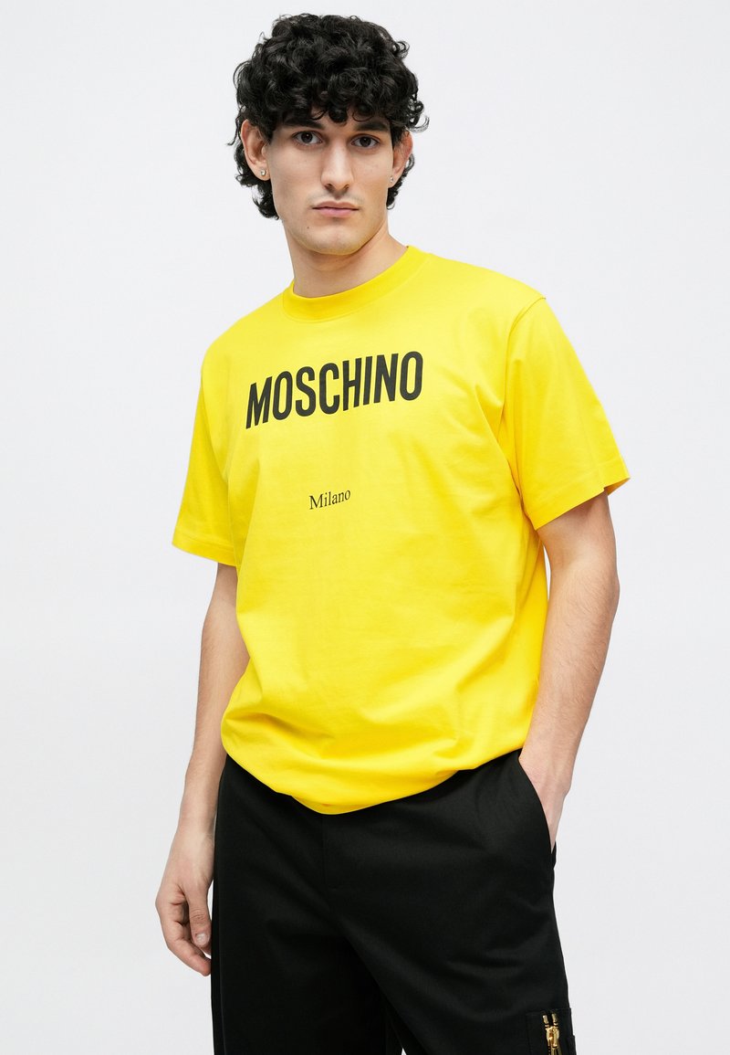 Gul bomuld T-shirt med en fremtrædende sort "MOSCHINO"-logo og et mindre "Milano"-print, korte ærmer og rund halsudskæring.