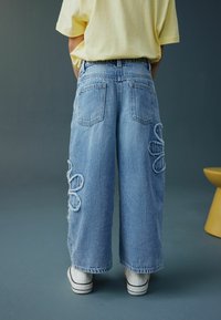 Šviesiai mėlynos plačios kojų džinsinės kelnės su apledėjusiomis gėlių raštais šone, su kišenėmis gale ir laisvu siluetu. Žemiau matomi balti sportbačiai.