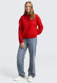 Czerwony sweter z dzianiny o okrągłym dekolcie, luźnym kroju i prążkowanych mankietach, w zestawieniu z jasnoniebieskimi jeansami o luźnym kroju i białymi tenisówkami.