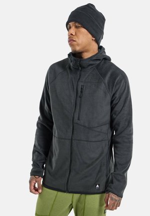 Schwarze Fleecejacke mit Reißverschluss, Kapuze und strukturierten Einsätzen. Mit einer Brusttasche mit Reißverschluss. Kombiniert mit grünen Hosen.
