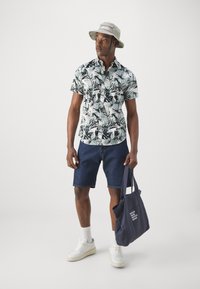 Camicia a maniche corte con bottoni, con un motivo tropicale di foglie in bianco e nero; abbinata a pantaloni shorts in denim, sneakers bianche e un cappello a secchiello grigio.