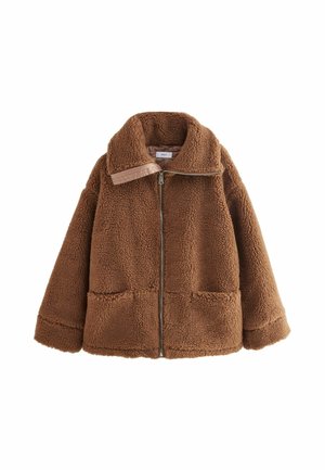 Veste marron en tissu duveteux avec fermeture éclair portée sur un haut blanc, mains dans les poches, associée à un jean patchwork bleu à l'extérieur près d'une rambarde en pierre.