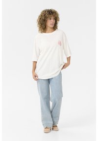 Valge ülev oversized T-särk, millel on roosa lille graafika vasakul rinnal, koos helesiniste laiade pükstega ja beeži libisevaäärisega kingadega.