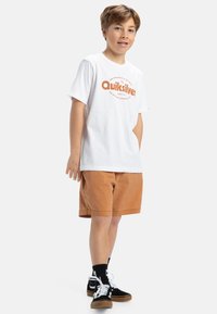Bílé tričko s krátkým rukávem a oranžovým logem "Quiksilver", v kombinaci s béžovými šortkami a černými teniskami s bílými tkaničkami a ponožkami.