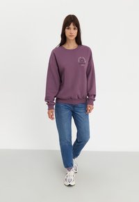 Paarse oversized sweatshirt met geribbelde manchetten en zoom; heeft een kleine grafische print op de borst. Gecombineerd met blauwe jeans en meerkleurige sneakers.