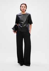 Top de couro preto com mangas curtas e decote redondo, combinado com calças largas pretas. O modelo segura uma carteira preta, posando contra um fundo neutro.