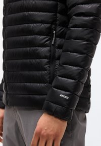 Μανίκι μαύρου καπιτονέ μπουφάν με εκτύπωση "800" κοντά στην μανσέτα, εμφανής τσέπη με φερμουάρ, φορεμένο πάνω από γκρι παντελόνια.