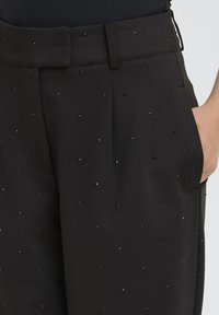 Primo piano di pantaloni neri con piccole decorazioni di perline lucide, vita alta con cintura in tessuto coordinato e tasca laterale che tiene una mano.