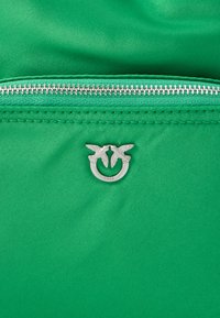 Tessuto verde con una superficie liscia presenta una cerniera argentata e un logo circolare con due uccelli. Le cuciture sono visibili lungo i bordi.