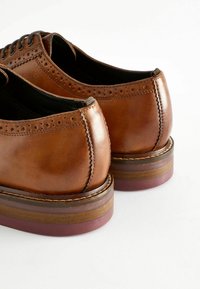 Brune lædersko med en glat finish, brogue-detaljer, en rund tå og en lagdelt træsål med en maroon accent.