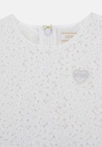 Robe en dentelle blanche avec des motifs floraux, un col rond et une fermeture éclair dans le dos. Comprend un logo "GUESS" en forme de cœur transparent.