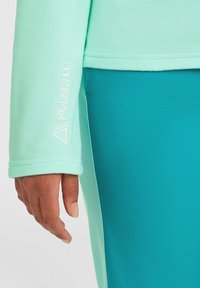Mintgrön fleecejacka ärm med en reflexande "Polartec"-logotyp. Åtföljd av en teal tyg, som visar en slät textur och åtsittande design.
