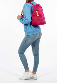 Magenta backpack with adjustable straps, front flap, yellow ribbon detail, and zipper accents, worn by a person in a blue sweatshirt and jeans.