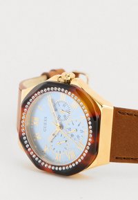 Orologio ottagonale in tonalità oro con cinturino in pelle marrone. Presenta un quadrante blu chiaro, numeri romani, tre sottomisure e decorazioni in cristallo.