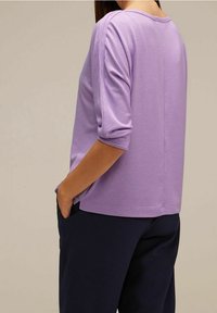 Lila lättviktig topp med trekvartslånga ärmar, rund halsringning och en avslappnad passform, kombinerad med mörkblå byxor.