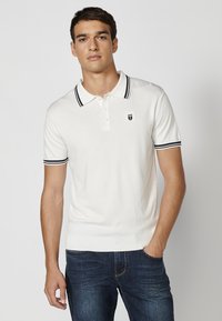 Biely polo tričko s krátkym rukávom a čiernymi pruhovanými golierom a manžetami, s malým logom na hrudi; hladká textúra, priliehavý strih.